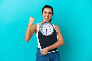 Mavi arka planda, tartı makinesiyle izole edilmiş beyaz bir kadın zafer jesti yapıyor.