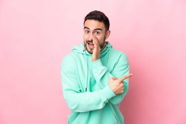 Pembe arka planda soyutlanmış beyaz tenli yakışıklı bir adam bir ürün sunmak ve bir şeyler fısıldamak için kenarı işaret ediyor.