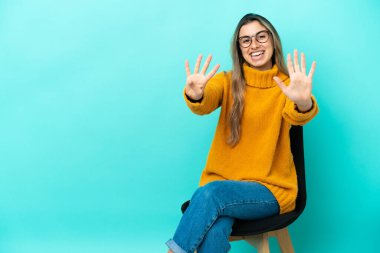 Mavi arka planda izole edilmiş bir sandalyede oturan beyaz bir kadın dokuz parmaklı sayıyor.