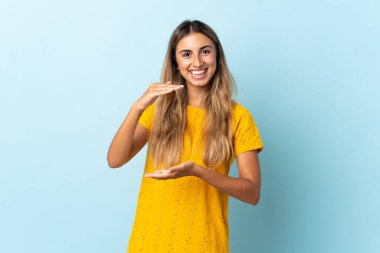 İzole edilmiş mavi arka plan üzerinde genç İspanyol kadın bir ilan vermek için avucunun içinde hayali bir şey tutuyor.