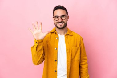 Pembe arka planda izole edilmiş beyaz, yakışıklı genç bir adam parmaklarıyla beşi sayıyor.