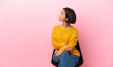 Genç melez kadın, arka planda pembe arka planda izole edilmiş bir sandalyede oturuyor ve arkasına bakıyor.