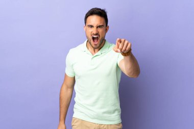 Mor arka planda izole edilmiş genç yakışıklı hayal kırıklığına uğramış ve ön tarafı işaret ediyor.