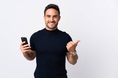 Beyaz arka planda izole edilmiş cep telefonu kullanan genç beyaz bir adam bir ürün sunmak için kenarı işaret ediyor