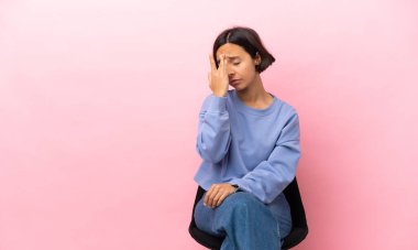 Genç melez kadın pembe arka planda izole edilmiş bir sandalyede oturuyor ve başı ağrıyor.