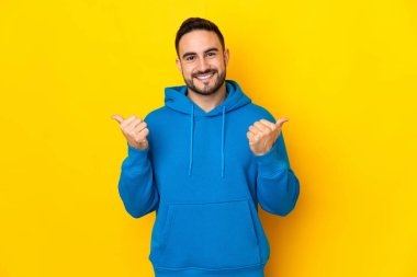 Sarı arka planda izole edilmiş beyaz, yakışıklı genç bir adam. Baş parmakları havada ve gülümsüyor.