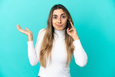 Mavi arka planda izole edilmiş cep telefonu kullanan genç beyaz kadın ellerini kaldırırken şüpheye düşüyor