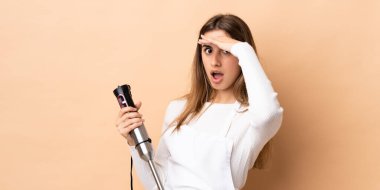 Arka planda el blenderi kullanan bir kadın yan tarafa bakarken sürpriz jest yapıyor.