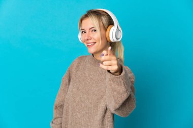 Genç Rus kadın mavi arka planda izole edilmiş müzik dinliyor ve ön tarafı işaret ediyor.