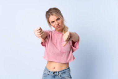 Sarışın kadın, izole edilmiş mavi arka planda baş parmağını iki eliyle gösteriyor.