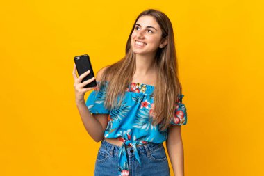 İzole edilmiş sarı arka planda cep telefonu kullanan genç bir kadın gülümserken yukarı bakıyor.
