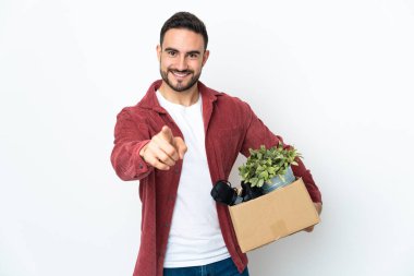 Beyaz arka planda izole edilmiş eşyalarla dolu bir kutuyu alıp mutlu bir ifadeyle ön tarafı işaret eden genç beyaz bir adam hamle yapıyor.