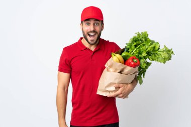 Sakallı genç bir adam elinde bir çanta dolusu sebzeyle beyaz arka planda şaşırmış bir yüz ifadesi var.