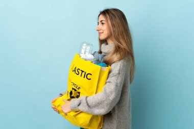 Geri dönüşüm için plastik şişelerle dolu bir torba tutarak yalıtılmış maviliğin üzerinde yan tarafa bakıyor.