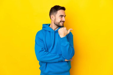 Sarı arka planda izole edilmiş beyaz, yakışıklı genç bir adam bir ürün sunmak için kenarı işaret ediyor.