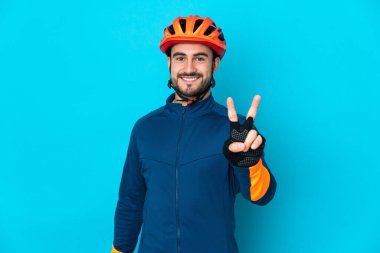 Mavi arka planda izole edilmiş genç bisikletçi gülümsüyor ve zafer işareti gösteriyor.