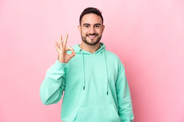 Pembe arka planda izole edilmiş beyaz, yakışıklı genç bir adam parmaklarıyla tamam işareti gösteriyor.