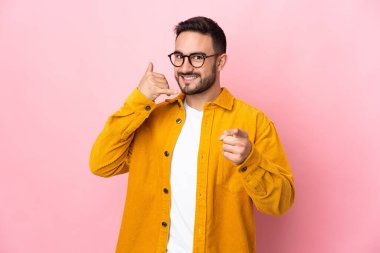 Beyaz tenli yakışıklı adam pembe arka planda izole edilmiş telefon hareketlerini yapıyor ve ön tarafı işaret ediyor.