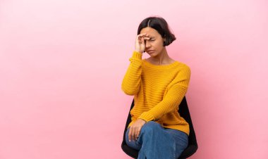 Genç melez kadın pembe arka planda izole edilmiş bir sandalyede oturuyor yorgun ve hasta bir ifadeyle.