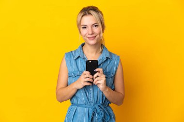 Genç Rus kadın sarı arka planda izole edilmiş kameraya bakıyor ve cep telefonunu kullanırken gülümsüyor.