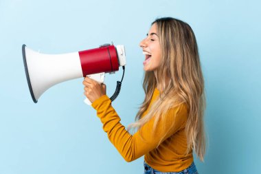 İzole edilmiş mavi arka planda genç İspanyol kadın megafondan bir şey duyurmak için bağırıyor.