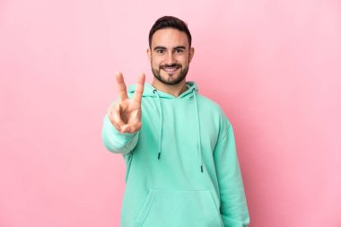 Beyaz tenli yakışıklı adam pembe arka planda izole edilmiş gülümsüyor ve zafer işareti gösteriyor.