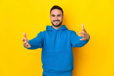 Sarı arka planda izole edilmiş beyaz, yakışıklı genç bir adam sunar ve davet eder.