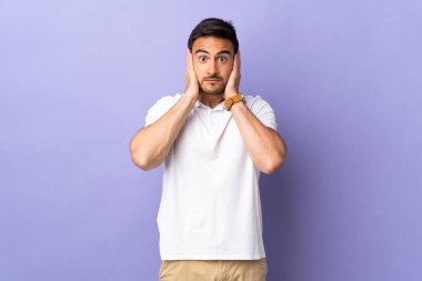 Mor arka planda izole edilmiş genç yakışıklı adam hayal kırıklığına uğramış ve kulaklarını kapatmış.