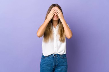 İzole edilmiş mor arka planı olan genç bir kadın gözlerini elleriyle kapatıyor.