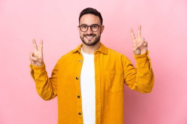 Pembe arka planda izole edilmiş beyaz, yakışıklı genç bir adam iki eliyle zafer işareti gösteriyor.