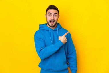 Sarı arka planda izole edilmiş beyaz, yakışıklı genç adam şaşırmış ve işaret ediyor.