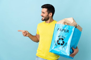 Mavi arka planda izole edilmiş bir geri dönüşüm çantası tutan genç beyaz adam bir ürünü sunmak için yan tarafı işaret ediyor.