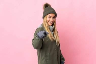 Pembe arka planda izole edilmiş, kış şapkalı genç kadın şaşırmış ve ön tarafı işaret ediyor.
