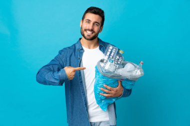 Mavi arka planda izole edilmiş plastik şişelerle dolu bir çanta taşıyan genç beyaz bir adam.