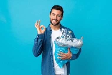 Mavi arka planda izole edilmiş plastik şişelerle dolu bir çanta taşıyan genç beyaz bir adam parmaklarıyla tamam işareti gösteriyor.