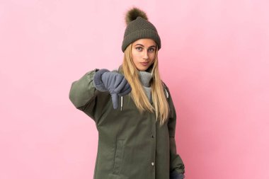 Pembe arka planda izole edilmiş, şapkalı genç bir kadın olumsuz bir ifadeyle baş parmağını aşağı gösteriyor.