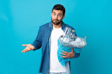 Mavi arka planda izole edilmiş plastik şişelerle dolu bir çanta taşıyan genç beyaz bir adam omuzlarını kaldırırken şüpheyle hareket ediyor.