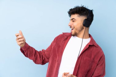 Arap yakışıklı adam mavi arka planda izole edilmiş müzik dinliyor ve gitar hareketi yapıyor.