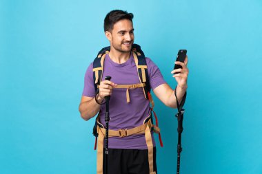Sırt çantası ve yürüyüş direkleri olan beyaz bir adam mavi arka planda izole edilmiş bir şekilde selfie çekiyor.