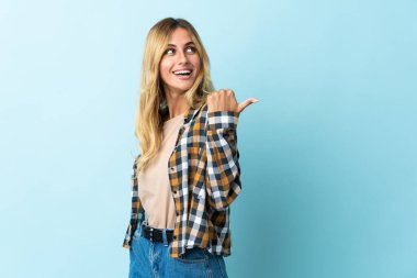 Mavi arka planda izole edilmiş genç sarışın Uruguaylı bir ürünü sunmak için kenarı işaret ediyor