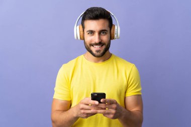 Mor arka planda sakallı genç yakışıklı bir adam cep telefonuyla müzik dinliyor ve ön tarafa bakıyor.