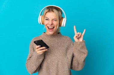 Genç Rus kadın mavi arka planda izole edilmiş, hareketli bir rock hareketi yaparak müzik dinliyor.