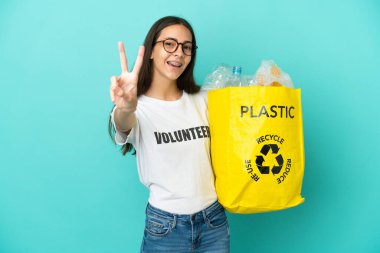 Gülümseyen ve zafer işareti gösteren plastik şişelerle dolu bir çanta tutan genç Fransız kız.