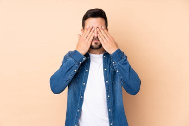 Beyaz tenli yakışıklı adam bej arka planda izole edilmiş, gözleri elle kapatılmış.