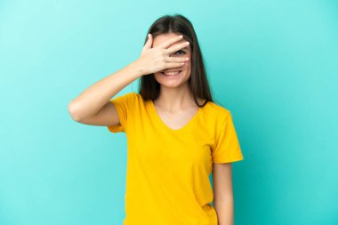Mavi arka planda izole edilmiş genç beyaz bir kadın. Gözleri elleriyle kaplıyor ve gülümsüyor.