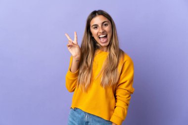İzole edilmiş mor arka planda genç İspanyol kadın gülümsüyor ve zafer işareti gösteriyor.