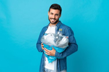 Mavi arka planda izole edilmiş ve mutlu bir ifadeyle izole edilmiş plastik şişelerle dolu bir çanta taşıyan genç beyaz bir adam.