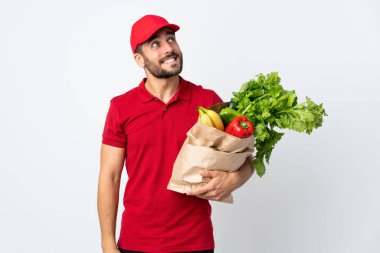 Sakallı genç adam elinde bir çanta dolusu sebzeyle beyaz arka planda izole edilmiş gülüyor ve yukarı bakıyordu.