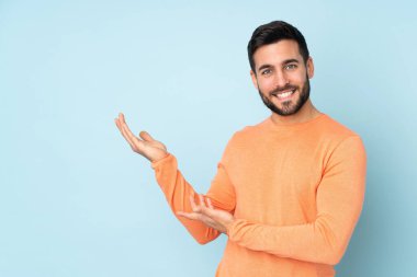 Beyaz tenli yakışıklı adam, izole edilmiş mavi arka plana davet edildiği için ellerini yana açıyor.