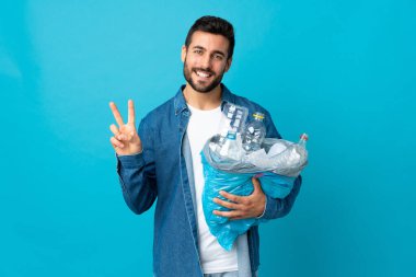Mavi arka planda izole edilmiş plastik şişelerle dolu bir çanta taşıyan genç beyaz adam gülümsüyor ve zafer işareti gösteriyor.